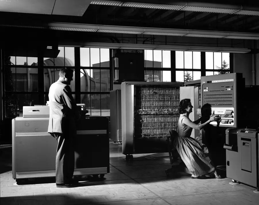 A man at a card reader and a woman at the console of an IBM 704 computer in 1958, in a hall lit by tall windows.